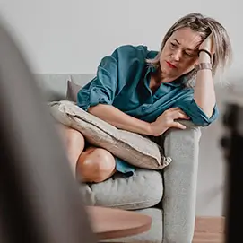 Femme assise sur un canapé, l'air pensif et préoccupé, tenant sa tête appuyée sur sa main. Elle semble plongée dans ses pensées, reflétant un moment de réflexion ou de fatigue.