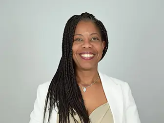 Portrait de Viviane Dupros souriante avec des tresses, portant une veste blanche, sur un fond neutre. Son expression reflète la confiance et la chaleur.