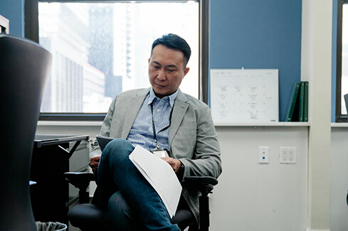 Homme d'affaires concentré, assis dans un bureau, tenant des documents et prenant des notes. Il est vêtu d'une veste grise et d'une chemise, avec un badge d'identification, et semble plongé dans son travail.