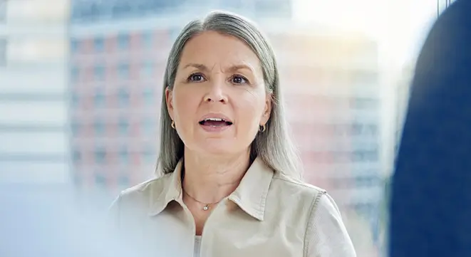 "Femme d'âge mûr, souriante et détendue, assise sur un canapé avec un smartphone à la main. Elle porte des lunettes et une chemise sombre, dans un intérieur moderne et accueillant avec des plantes en arrière-plan."